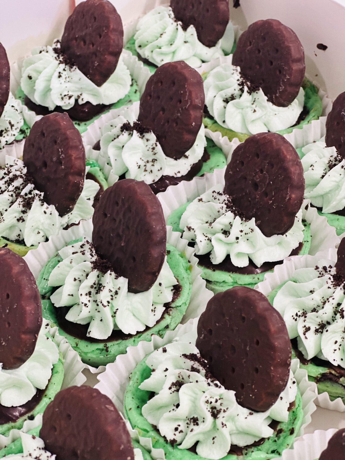 IMG_8768 A dozen cupcakes decorated with mint green frosting, topped with chocolate cookies partially embedded in the frosting. The frosting appears swirled and sprinkled with chocolate crumbs. The cupcakes are arranged in white paper liners.